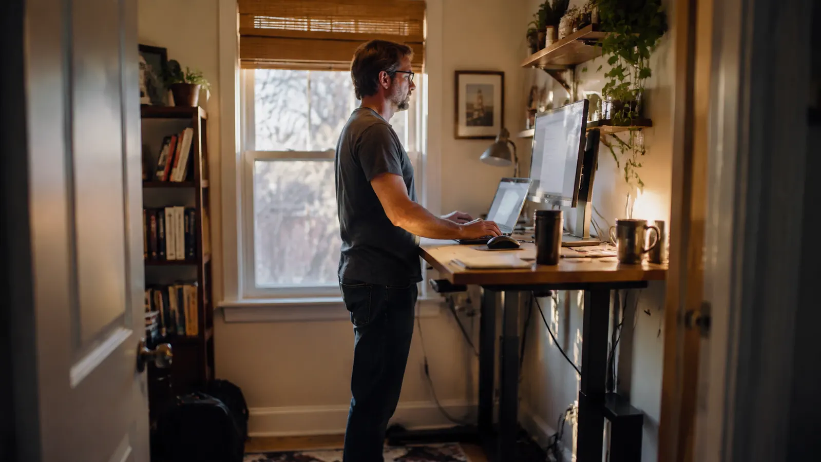 A spare bedroom home office in the late afternoon, standing desk raised to standing height with a laptop and monitor, soft window light