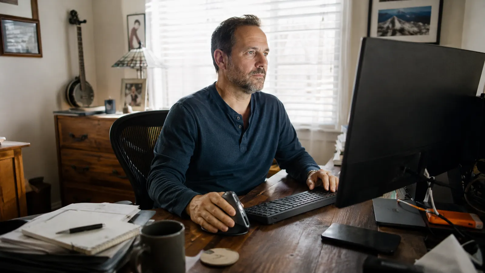 A man's hand resting on a vertical ergonomic mouse beside a keyboard on a home desk in warm afternoon light